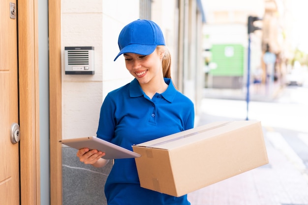 Young delivery woman at outdoors holding boxes and a tablet with happy expression