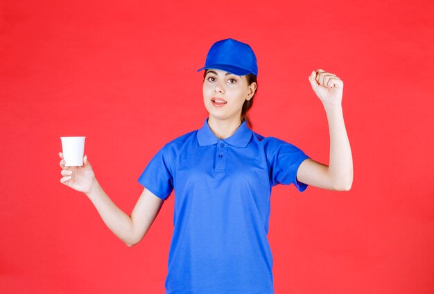 Young deliverywoman in blue cap posing with plastic cup of tea on red.