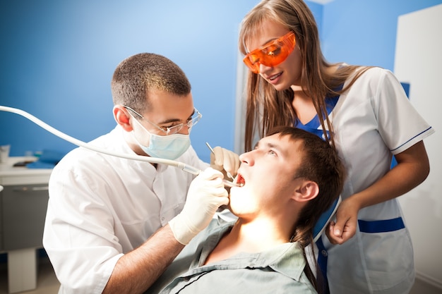 Young dentist doctor man in white uniform and special mask and young nurse woman examining man patient in dental office