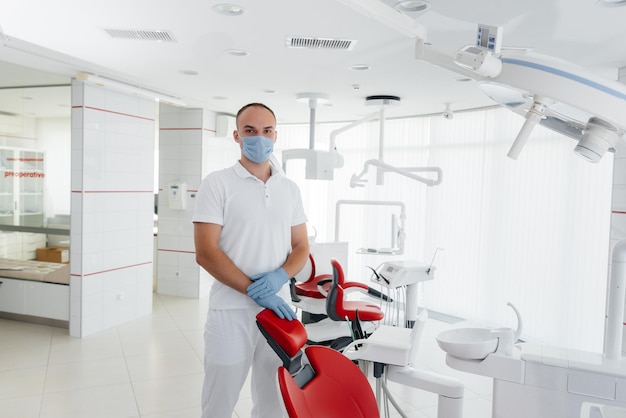 A young dentist in a mask stands near a red dental chair and smiles in modern white dentistry Modern dentistry and prosthetics