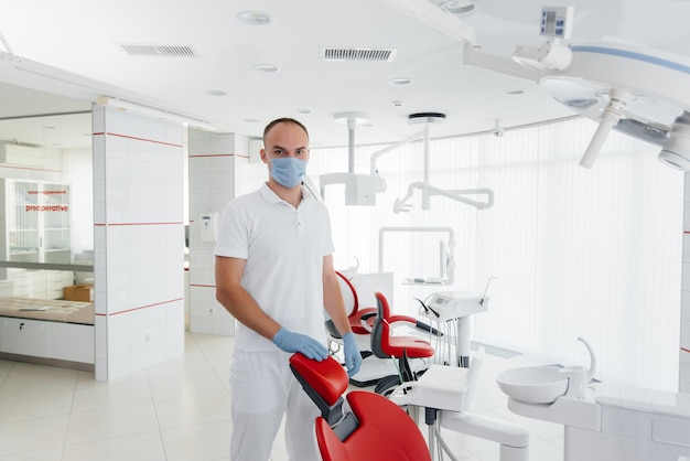 A young dentist in a mask stands near a red dental chair and smiles in modern white dentistry Modern dentistry and prosthetics