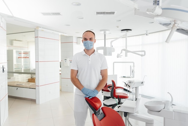 A young dentist in a mask stands near a red dental chair and smiles in modern white dentistry Modern dentistry and prosthetics