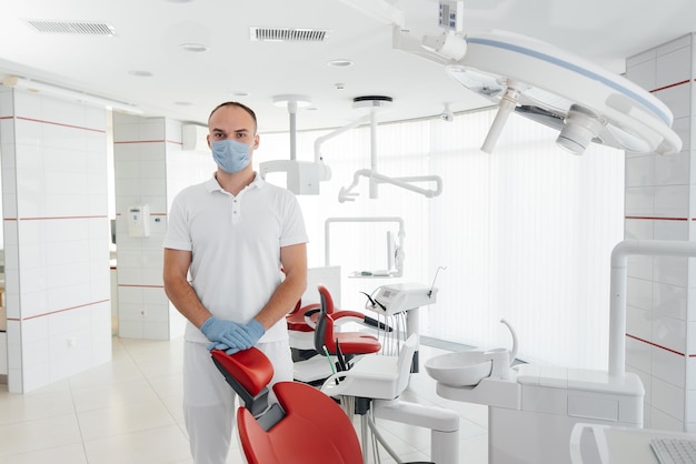 A young dentist in a mask stands near a red dental chair and smiles in modern white dentistry Modern dentistry and prosthetics