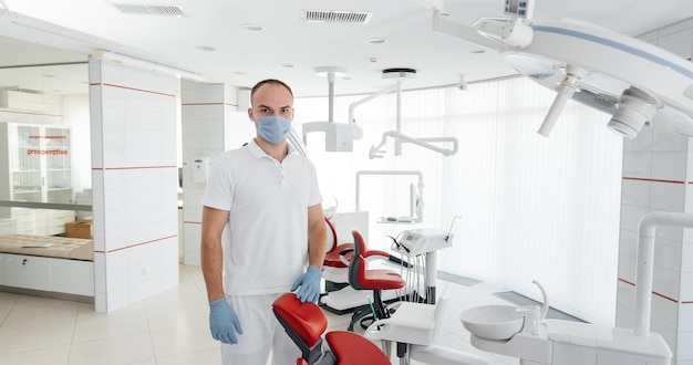 A young dentist in a mask stands near a red dental chair and smiles in modern white dentistry Modern dentistry and prosthetics