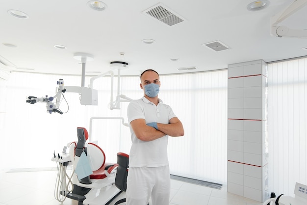 A young dentist in a mask stands near a red dental chair and smiles in modern white dentistry Modern dentistry and prosthetics