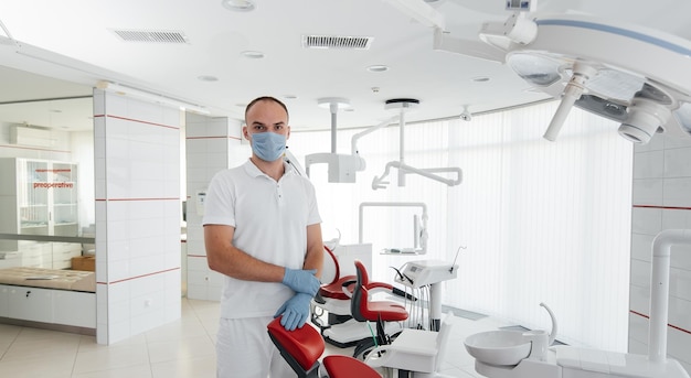 A young dentist in a mask stands near a red dental chair and smiles in modern white dentistry Modern dentistry and prosthetics