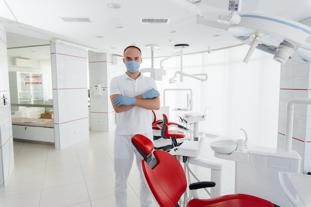 A young dentist in a mask stands near a red dental chair and smiles in modern white dentistry Modern dentistry and prosthetics