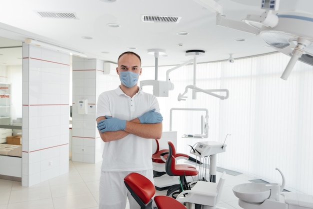 A young dentist in a mask stands near a red dental chair and smiles in modern white dentistry Modern dentistry and prosthetics