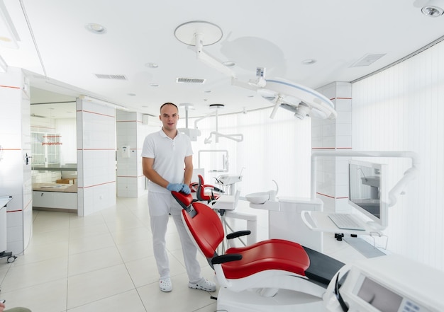 A young dentist stands near a red dental chair and smiles in modern white dentistry Treatment and prevention of caries from youth Modern dentistry and prosthetics