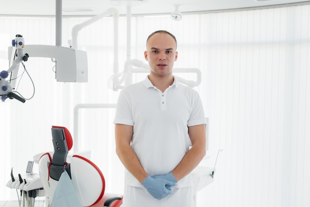 A young dentist stands near a red dental chair and smiles in modern white dentistry Treatment and prevention of caries from youth Modern dentistry and prosthetics