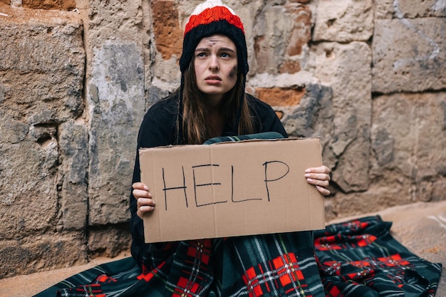 A young depressed homeless girl or woman sitting alone on a fence under a keep wall outside in cold weather she asks for help