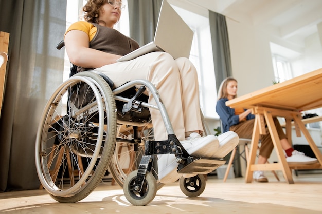 Young disable businesswoman with laptop on knees sitting in wheelchair in office and surfing in the net against her colleague working by table