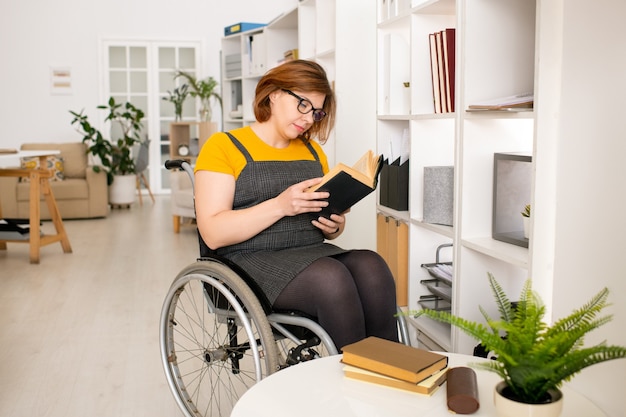 Young disable female in casualwear and eyeglasses sitting on wheelchair by shelf and reading book while getting home education