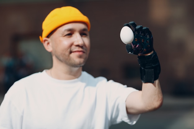 Young disabled man holds fragile chicken egg with artificial prosthetic hand in casual clothes