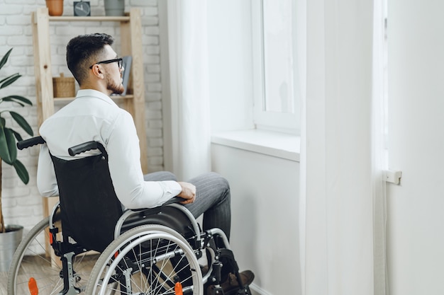 Young disabled man sitting in a wheelchair near the window at home