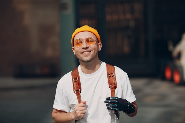 Young disabled man with artificial prosthetic hand in casual clothes and backpack walking at sunny c...