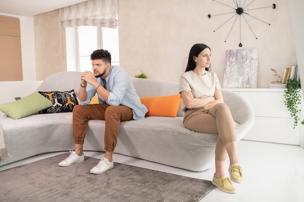 Young dissatisfied brunette female crossing arms by chest while sitting on couch in living-room against her pensive or offended husband