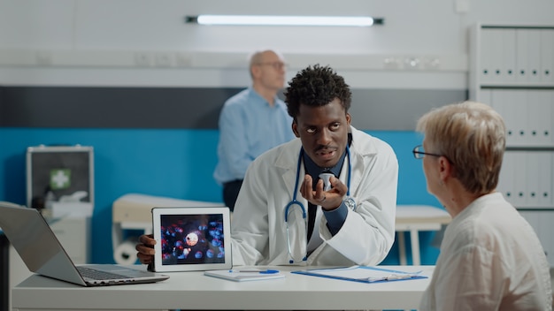 Young doctor analyzing virus animation on tablet with old woman at desk in medical cabinet. Medic and elderly patient looking at modern device showing coronavirus bacteria and danger