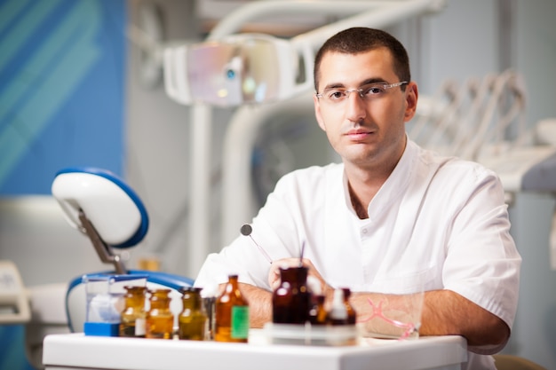 Young doctor dentist man in uniform sitting near dental chair in dental office in clinic with equipment at background