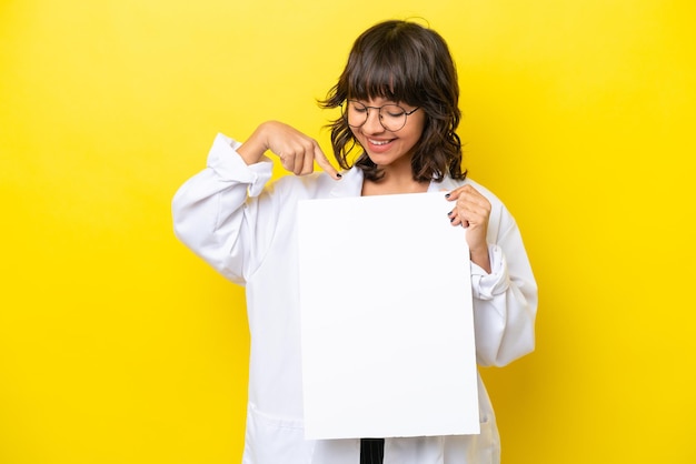 Young doctor latin woman isolated on yellow background holding an empty placard with happy expression and pointing it