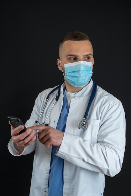 Young doctor man in medical uniform and mask stethoscope hold mobile cell phone isolated on black