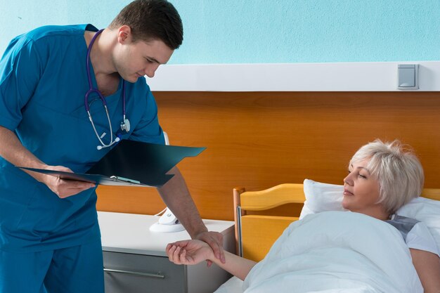 Young doctor in uniform with phonendoscope on his neck is measuring the pulse of his female patient, who is lying in the hospital bed in the hospital ward. Healthcare concept