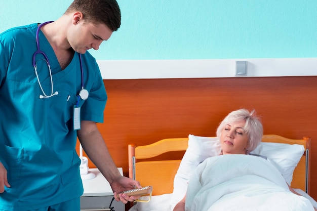 Young doctor in uniform with phonendoscope on his neck regulates a hospital bed of his female patient using a special console in the hospital ward. Healthcare concept