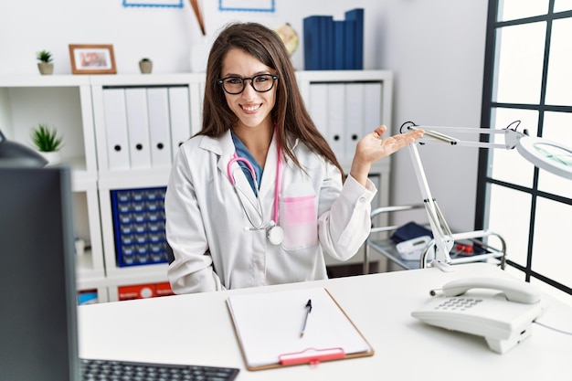 Young doctor woman wearing doctor uniform and stethoscope at the clinic smiling cheerful presenting and pointing with palm of hand looking at the camera