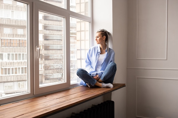 Young dreamy girl with dreadlocks sitting on a window