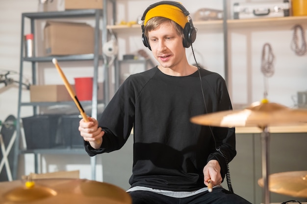 Photo young drummer with headphones holding drumsticks over cymbals while going to beat one of them during individual rehearsal