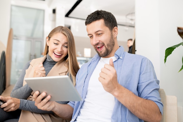 Young ecstatic students in casualwear looking at stuff on touchpad display and expressing excitement