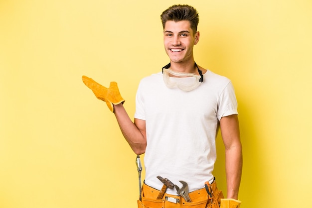 Young electrician caucasian man isolated on yellow background showing a copy space on a palm and holding another hand on waist.