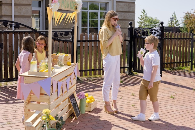 Young elegant blond woman and her little son drinking fresh homemade lemonade while standing by wooden stall with two girls