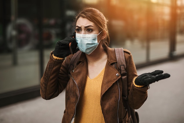 Young and elegant business woman walking on empty city street. She using phone while wearing protective mask to protect herself from dangerous virus. Corona virus or Covid-19 concept.