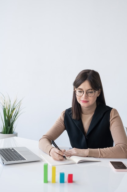 Young elegant female broker looking at multi-color cube chart on desk while analyzing company finances by workplace