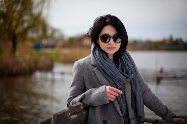 Young elegant girl in black glasses, a gray coat and a gray scarf on a walk in the park by the lake