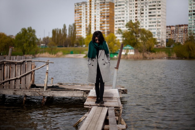 Young elegant girl in black glasses, a gray coat and a green scarf on a walk in the park by the lake