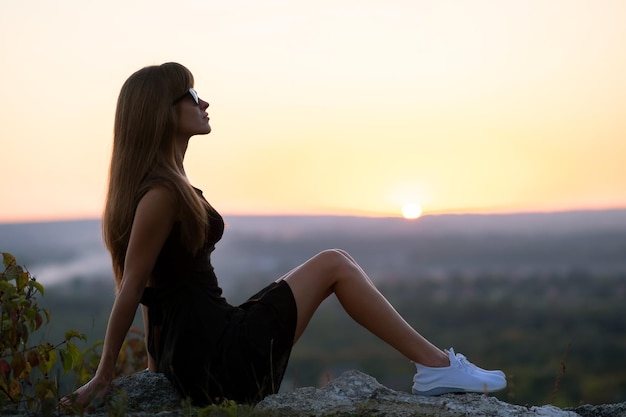 Young elegant woman in black short dress and white sneaker shoes sitting on a rock relaxing outdoors at summer evening Fashionable lady enjoying warm sunset in nature