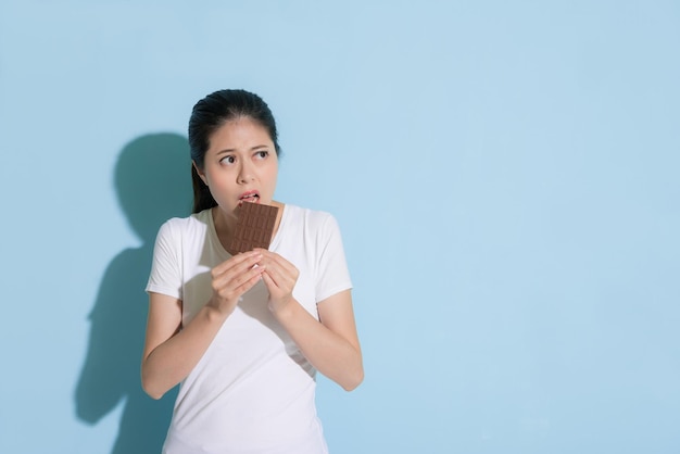 young elegant woman eating sweet chocolate dessert food in blue background and looking at empty area worry about tooth healthy.