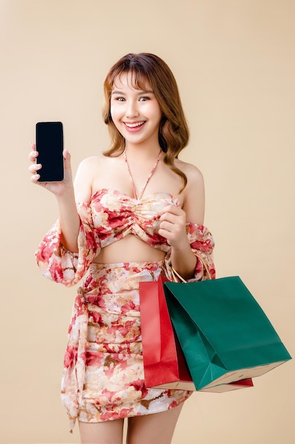 Young energetic Asian woman holding blank use smartphone searching shop retail with colorful shopping bags on beige background Online shoping futuristic concept