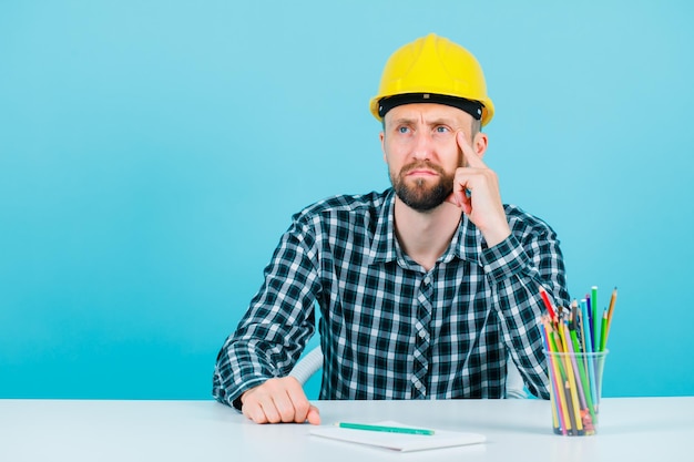 Young engineer man is looking away by holding hand on temple on blue background