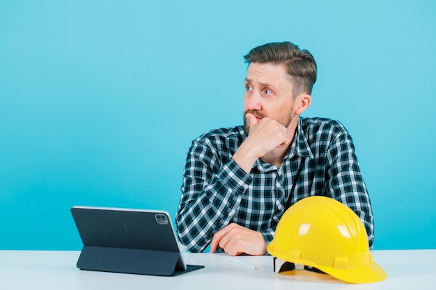 Young engineer man is thinking by holding thumb on lips and sitting in front of tablet on blue background