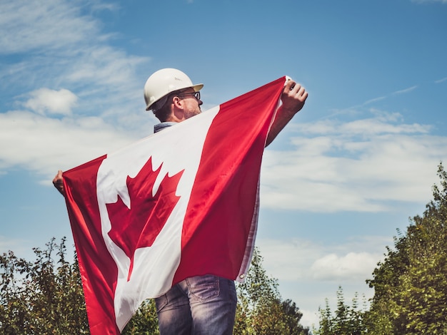 Young engineer, white hardhat and Canadian Flag in the park against the backdrop of green trees and the setting sun, looking into the distance. Close-up. Concept of labor and employment