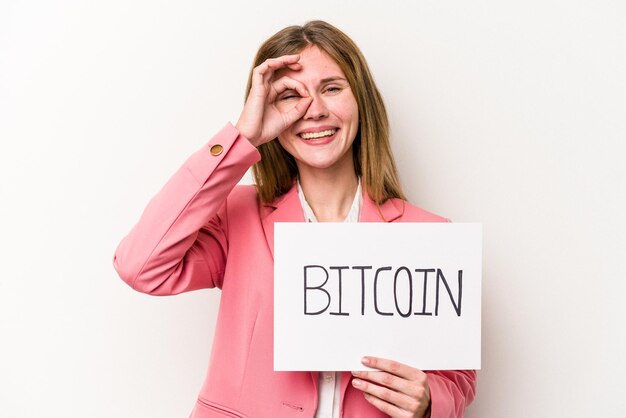 Young English business woman holding a bitcoin placard isolated on white background excited keeping ok gesture on eye