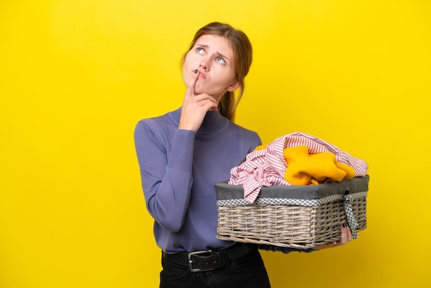 Young English woman holding a clothes basket isolated on yellow background having doubts while looking up