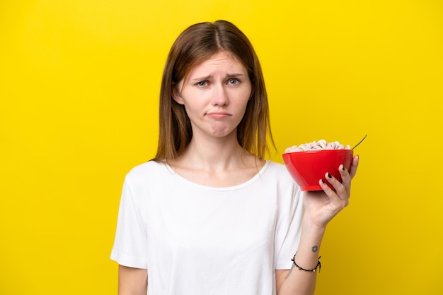 Young English woman holding cup of coffee isolated on yellow background with sad expression