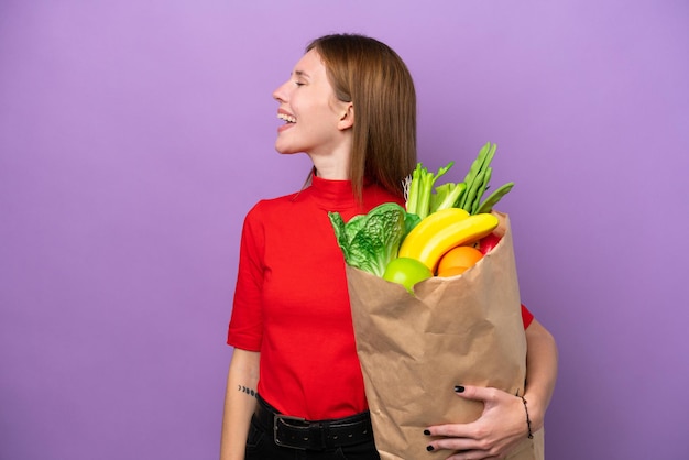 Young English woman holding a grocery shopping bag isolated on purple background laughing in lateral position