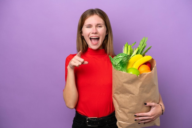 Young English woman holding a grocery shopping bag isolated on purple background surprised and pointing front