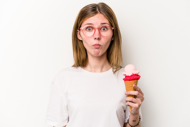 Young English woman holding an ice cream isolated on white background shrugs shoulders and open eyes confused