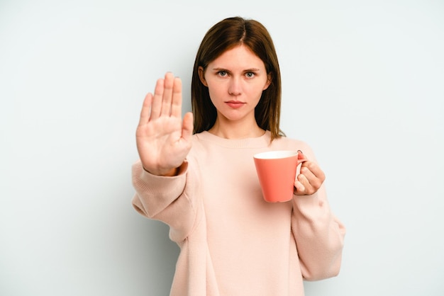 Young English woman holding a mug isolated on blue background standing with outstretched hand showing stop sign, preventing you.